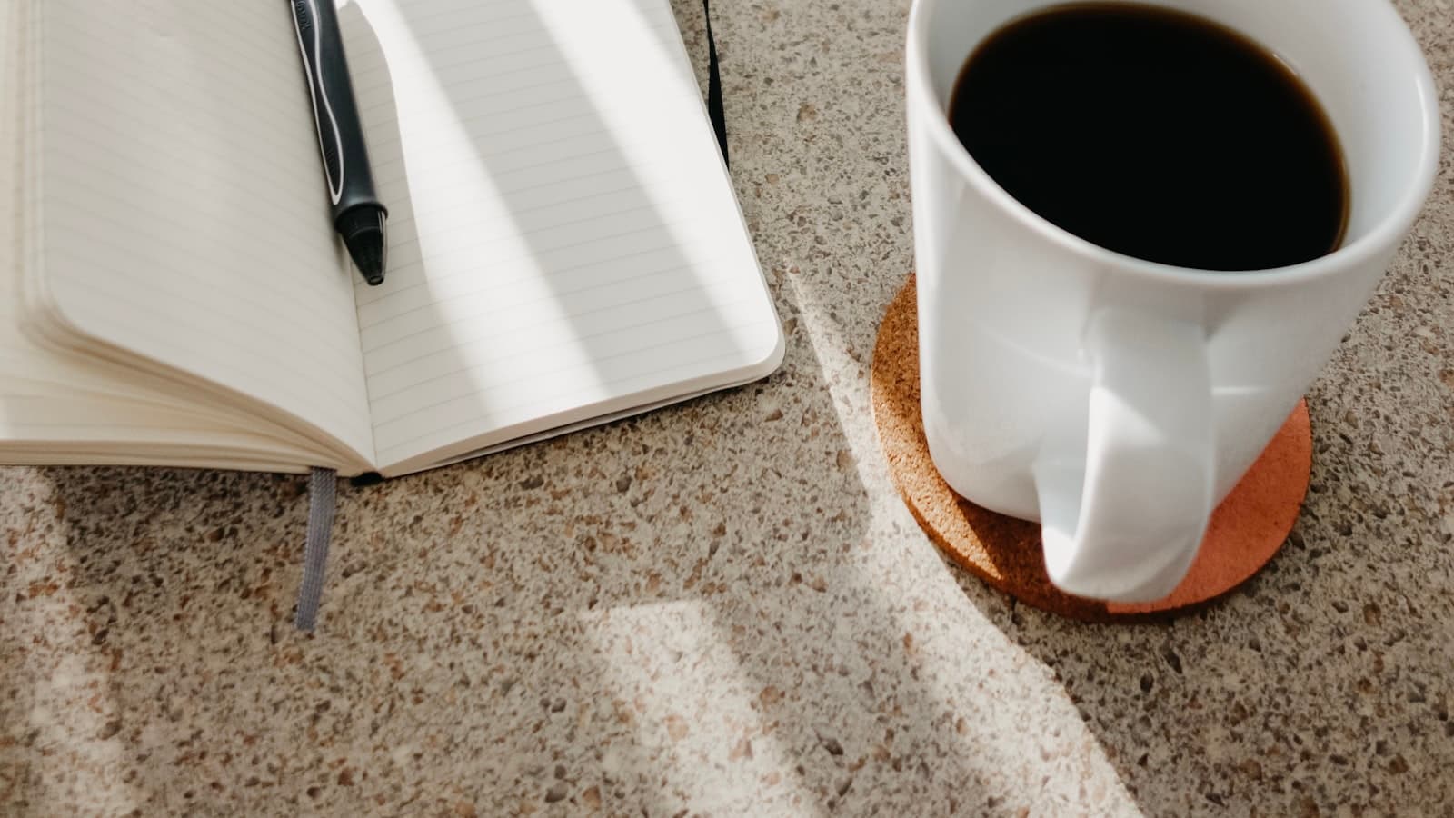 An open notebook and pen beside a mug of coffee on a kitchen counter