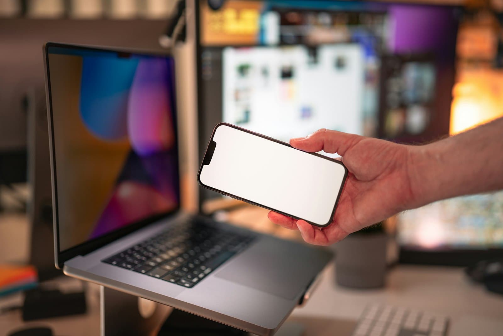 A hand holding a phone up in front of a laptop on a desk